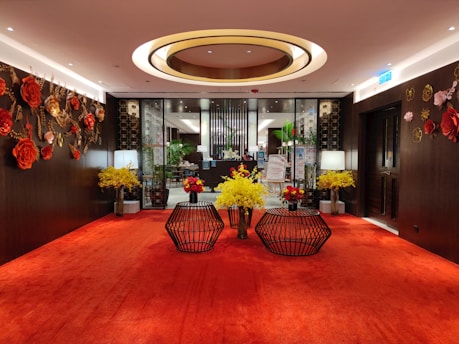 A luxurious interior space with a red carpet floor and elaborate floral decorations on the walls. Two black wireframe tables in the center hold vibrant yellow and red floral arrangements. The wall decor features large red and orange flowers. Two lamps with white shades are placed symmetrically near the back wall, which is made of dark wood. The ceiling has a circular design with warm lighting, enhancing the ambiance.