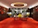 A luxurious interior space with a red carpet floor and elaborate floral decorations on the walls. Two black wireframe tables in the center hold vibrant yellow and red floral arrangements. The wall decor features large red and orange flowers. Two lamps with white shades are placed symmetrically near the back wall, which is made of dark wood. The ceiling has a circular design with warm lighting, enhancing the ambiance.