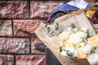 A close-up of a person wearing a graduation gown holding a bouquet wrapped in newspaper. The bouquet is composed of light-colored roses and greenery. The brick wall background provides a textured contrast to the subject.