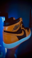 A high-top sneaker with a combination of black and gold leather, featuring a white sole. The iconic wing logo is visible on the side of the shoe. The background is blurred, highlighting the shoe in the foreground.
