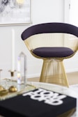 A modern chair with a golden wireframe base and dark cushion is positioned in a stylish and minimalistic room. A glass coffee table holds decorative items and a book with bold text. A framed abstract artwork with floral patterns hangs on the white wall, contributing to the contemporary aesthetic.