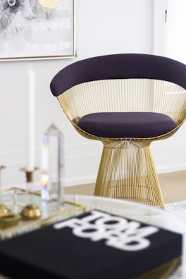 A modern chair with a golden wireframe base and dark cushion is positioned in a stylish and minimalistic room. A glass coffee table holds decorative items and a book with bold text. A framed abstract artwork with floral patterns hangs on the white wall, contributing to the contemporary aesthetic.