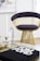 A modern chair with a golden wireframe base and dark cushion is positioned in a stylish and minimalistic room. A glass coffee table holds decorative items and a book with bold text. A framed abstract artwork with floral patterns hangs on the white wall, contributing to the contemporary aesthetic.