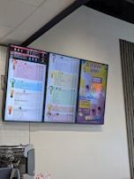 Digital menu boards display a selection of beverages such as milk tea, yogurt, espresso, and specialty drinks. The menu is colorful, showing prices and options in various fonts and colors. A prominent section advertises a 'Blueberry Series' with vibrant visuals of lemons and blueberries. Below the screens, a coffee machine and some promotional items are visible.