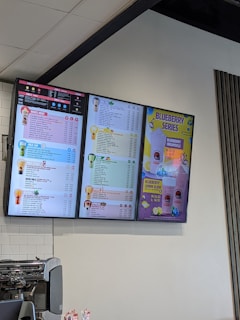 Digital menu boards display a selection of beverages such as milk tea, yogurt, espresso, and specialty drinks. The menu is colorful, showing prices and options in various fonts and colors. A prominent section advertises a 'Blueberry Series' with vibrant visuals of lemons and blueberries. Below the screens, a coffee machine and some promotional items are visible.