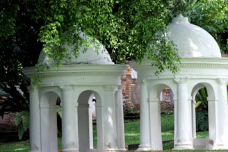Two white neoclassical structures with domed tops stand amidst lush green foliage. The architecture features arches and detailed cornices, with a partially visible brick wall in the background. The surrounding trees and plants create a serene mix of nature and historical design.