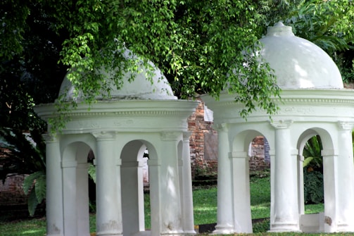 Two white neoclassical structures with domed tops stand amidst lush green foliage. The architecture features arches and detailed cornices, with a partially visible brick wall in the background. The surrounding trees and plants create a serene mix of nature and historical design.