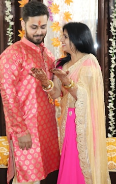 A couple is dressed in traditional attire, standing close together and smiling warmly at each other. The man is wearing a pink and gold patterned kurta while the woman is wearing a pink and gold saree. There is intricate henna on her hands. The background is adorned with yellow and white floral decorations.
