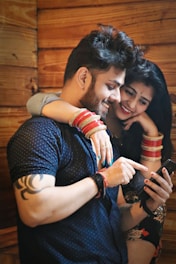 A couple is closely interacting with a smartphone. The man is pointing at the screen while the woman, wearing colorful bracelets, gently rests her arm over his shoulders, both smiling and engaged.