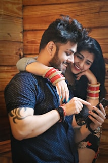 A couple is closely interacting with a smartphone. The man is pointing at the screen while the woman, wearing colorful bracelets, gently rests her arm over his shoulders, both smiling and engaged.