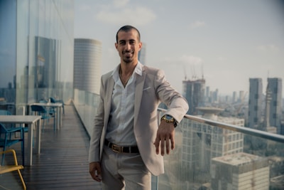 A man in a light-colored suit stands confidently on a rooftop terrace, overlooking a cityscape. There are tables and chairs around him, and modern skyscrapers can be seen in the background. The sky is clear with a few clouds, capturing a calm afternoon ambiance.