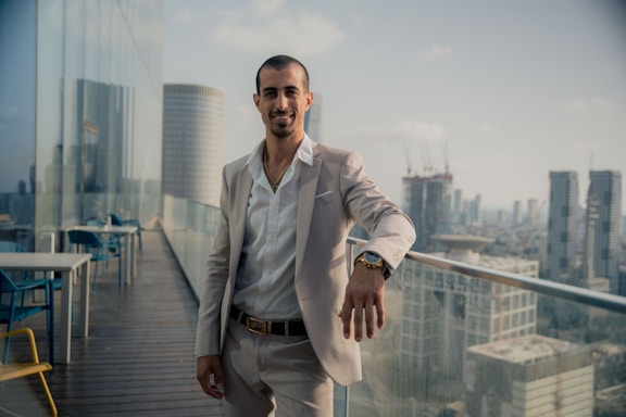 A man in a light-colored suit stands confidently on a rooftop terrace, overlooking a cityscape. There are tables and chairs around him, and modern skyscrapers can be seen in the background. The sky is clear with a few clouds, capturing a calm afternoon ambiance.