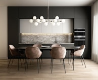 A modern kitchen and dining area with a sleek design. The space features a dark grey cabinet wall, white upper cabinets, and a marble backsplash. A large dining table is surrounded by six beige upholstered chairs with black metal legs. Above the table hangs a contemporary chandelier made of spherical lights. The flooring is a light wood, contributing to a warm and inviting atmosphere.