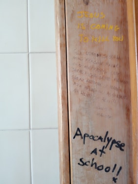 A wooden panel with two distinct writings in different styles and colors. The top text is in bold yellow, reading 'JESUS IS COMING TO KILL YOU.' Below are faint sentences in a smaller, cursive style, difficult to decipher. The bottom text is in black, bold handwriting, stating 'Apocalypse at school!!' The background consists of white square tiles.