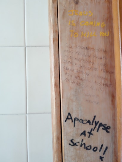 A wooden panel with two distinct writings in different styles and colors. The top text is in bold yellow, reading 'JESUS IS COMING TO KILL YOU.' Below are faint sentences in a smaller, cursive style, difficult to decipher. The bottom text is in black, bold handwriting, stating 'Apocalypse at school!!' The background consists of white square tiles.