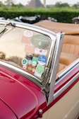 A vintage red convertible car displays various car show and rally stickers on its windshield. The car has a sleek chrome trim and tan leather interior. The background features blurred greenery and other classic cars.