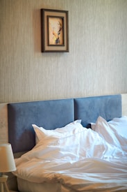 A neatly made bed with white sheets and pillows is positioned against a beige textured wall. Above the bed, a framed picture of a deer with antlers is centered on the wall. A bedside lamp is visible in the lower left corner.