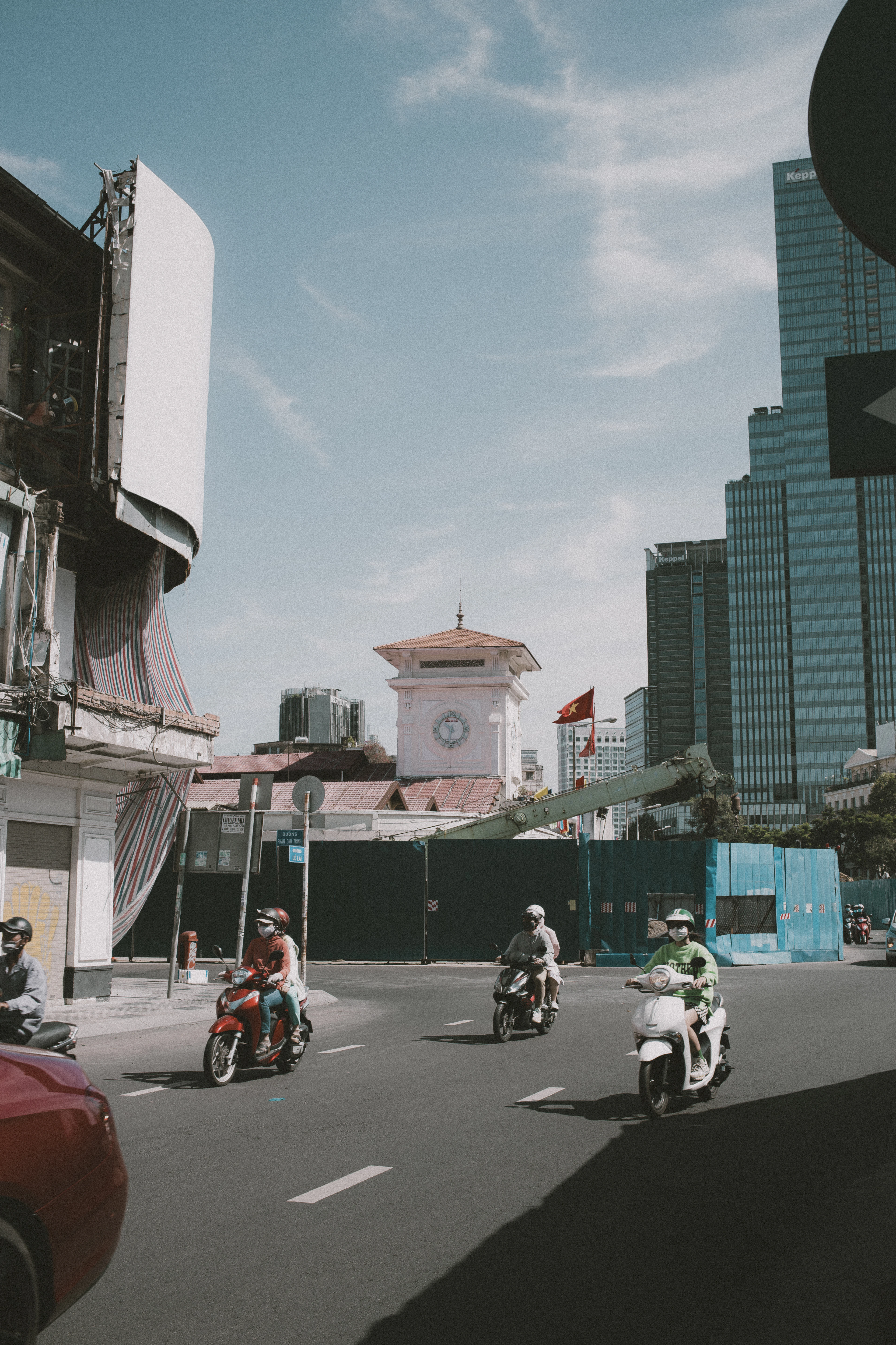 Motorbikes ride past Ben Thanh Market's clock tower with the Bitexco Financial Tower rising behind it under a clear sky.