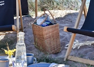 A wicker basket filled with snacks and chips sits on sandy terrain surrounded by tall grass. Nearby, there are two wooden and fabric deck chairs. A bottle of water and cups are placed on a table with a floral decoration.