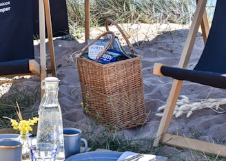A wicker basket filled with snacks and chips sits on sandy terrain surrounded by tall grass. Nearby, there are two wooden and fabric deck chairs. A bottle of water and cups are placed on a table with a floral decoration.