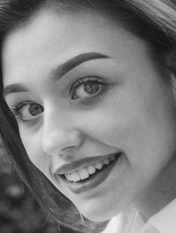 A close-up black and white portrait of a person smiling, showcasing well-defined eyes, eyebrows, and teeth. The lighting emphasizes the facial features against a slightly blurred background.