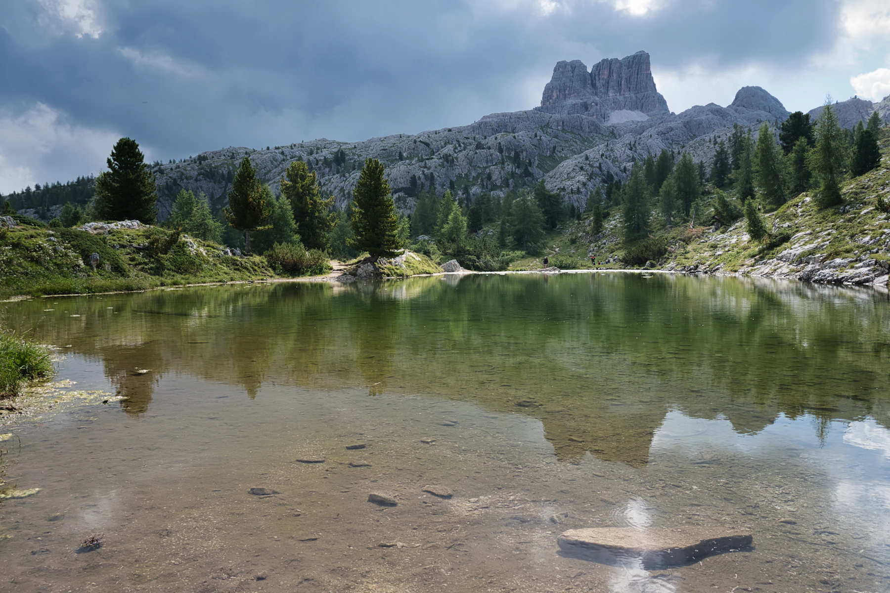 Photograph of A pale Dolomites summit reflects in a small alpine lake beneath a cloud-edged sky.