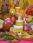 A vibrant arrangement featuring skincare products surrounded by fresh fruits and flowers. A jar of sea buckthorn cream and turmeric MCT scrub are prominently displayed among other products like hibiscus seed oil and pomegranate seed oil. Fruits such as a lemon, kiwi, coconut, and melon are artistically placed, alongside orange and pink blossoms, creating a lively and organic setting.