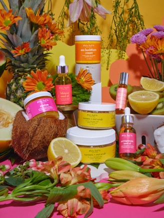 A vibrant arrangement featuring skincare products surrounded by fresh fruits and flowers. A jar of sea buckthorn cream and turmeric MCT scrub are prominently displayed among other products like hibiscus seed oil and pomegranate seed oil. Fruits such as a lemon, kiwi, coconut, and melon are artistically placed, alongside orange and pink blossoms, creating a lively and organic setting.