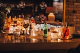 A cozy bar setting with a polished counter lined with various drinks and glasses. Soft, warm lighting creates an inviting atmosphere, reflecting off glass bottles and candles. Green glass bottle, orange cocktail garnished with a slice of fruit, and dim candlelight enhance the ambient mood.