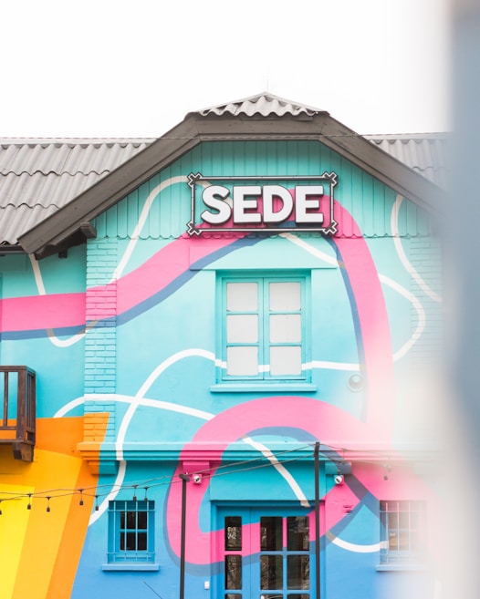 A building with vibrant, colorful street art features a prominent sign with the word 'SEDE'. The facade is painted in bright blue, pink, and orange with abstract, flowing patterns. There are windows and string lights hanging across the front.