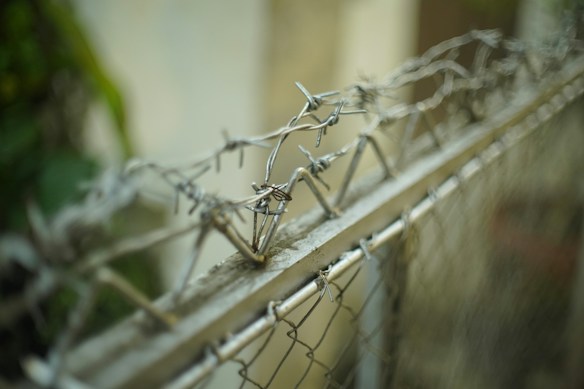 A close-up of barbed wire atop a chain-link fence, with the background softly blurred. The focus is on the sharp, twisted metal of the barbed wire, which runs horizontally along the top of the image.