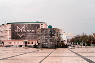 A large building with a banner in Cyrillic lettering occupies the left side of a spacious square. In the foreground, an object covered with scaffolding is visible. In the background, a church with golden domes can be seen. There are a few cars and pedestrians on the street, and trees line the road leading to the structure.