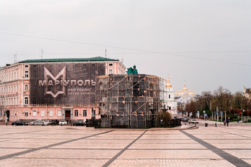 A large building with a banner in Cyrillic lettering occupies the left side of a spacious square. In the foreground, an object covered with scaffolding is visible. In the background, a church with golden domes can be seen. There are a few cars and pedestrians on the street, and trees line the road leading to the structure.