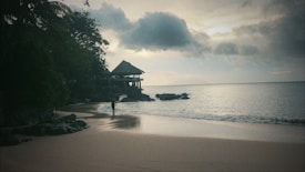 A serene beach setting during either dawn or dusk, with calm waves lapping against the shore. A lone figure walks along the sands, and a thatched-roof structure sits on the rocky outcrop by the water. The sky is overcast with moody clouds, reflecting a subdued light onto the ocean.