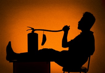 A silhouette of a person sitting on a chair with their feet up on a table, holding a hose connected to a fire extinguisher. The background is a gradient of deep orange tones.