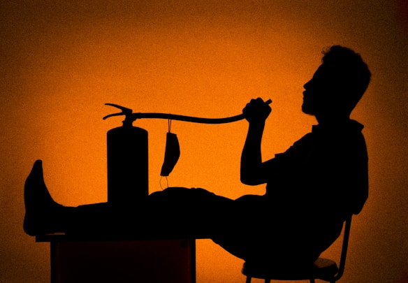 A silhouette of a person sitting on a chair with their feet up on a table, holding a hose connected to a fire extinguisher. The background is a gradient of deep orange tones.