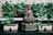 A serene Buddha statue sits at the center of a lush arrangement of potted green plants. The statue is holding a spherical object and is surrounded by decorative boxes with tassels on them. The setting is peaceful and elegantly adorned with a carved backdrop.