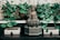 A serene Buddha statue sits at the center of a lush arrangement of potted green plants. The statue is holding a spherical object and is surrounded by decorative boxes with tassels on them. The setting is peaceful and elegantly adorned with a carved backdrop.