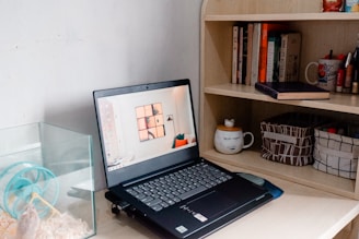 A small workspace featuring a laptop on a wooden desk next to a bookshelf. To the left, there's a clear cage with a small animal, possibly a hamster, and a wheel. The bookshelf contains books, mugs, and containers organized neatly. The laptop displays a home interior on its screen.