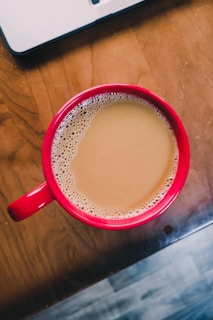 A red cup filled with coffee rests on a wooden table, with a laptop partially visible in the top left corner. The coffee is light brown and frothy near the edges, suggesting it may contain milk or cream.