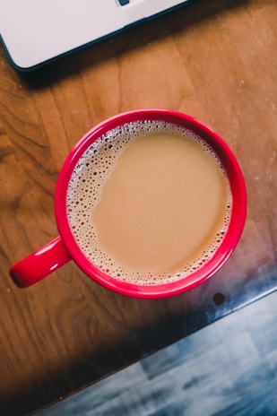 A red cup filled with coffee rests on a wooden table, with a laptop partially visible in the top left corner. The coffee is light brown and frothy near the edges, suggesting it may contain milk or cream.