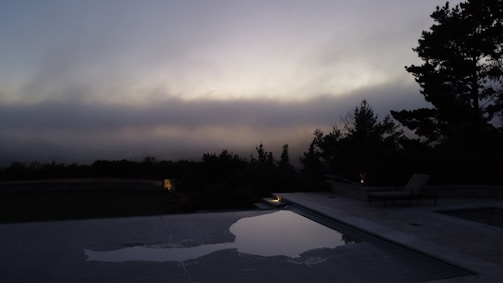 An outdoor pool scene at dusk, lights reflecting softly on the water surrounded by lush greenery.