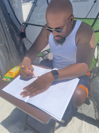 A person with sunglasses and a tank top is coloring in a sketchbook on a table, under a shaded tent. A set of colored pencils is placed beside the sketchbook.