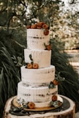 A three-tiered semi-naked cake adorned with white icing drips. The cake is decorated with small doughnuts and sprigs of green leaves, set against a natural, rustic background. It rests on a wooden slab, enhancing the outdoor, earthy theme.