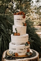 A three-tiered semi-naked cake adorned with white icing drips. The cake is decorated with small doughnuts and sprigs of green leaves, set against a natural, rustic background. It rests on a wooden slab, enhancing the outdoor, earthy theme.