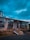 A construction site with a partially built structure made of bricks and wooden supports. The sky is cloudy, casting a dramatic light over the scene. Construction materials such as metal beams and wooden planks are scattered on the ground.
