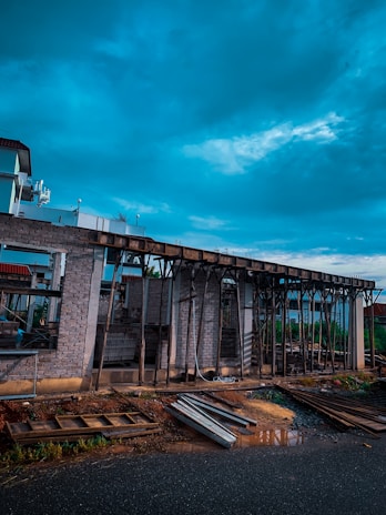 A construction site with a partially built structure made of bricks and wooden supports. The sky is cloudy, casting a dramatic light over the scene. Construction materials such as metal beams and wooden planks are scattered on the ground.