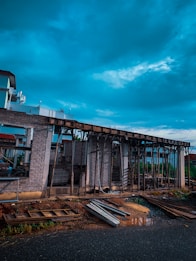 A construction site with a partially built structure made of bricks and wooden supports. The sky is cloudy, casting a dramatic light over the scene. Construction materials such as metal beams and wooden planks are scattered on the ground.