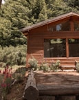 A wooden cabin with large windows is surrounded by lush green trees and colorful flowers. The wooden structure blends naturally with the forest scenery, adding to a rustic and peaceful environment.