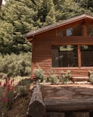 A wooden cabin with large windows is surrounded by lush green trees and colorful flowers. The wooden structure blends naturally with the forest scenery, adding to a rustic and peaceful environment.